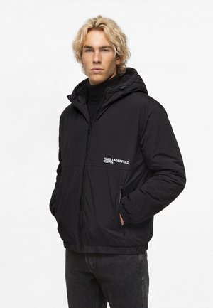 Blond mand iført en sort hættetrøje med "Karl Lagerfeld Jeans" logo, stående med hænderne i lommerne mod en ensfarvet hvid baggrund.