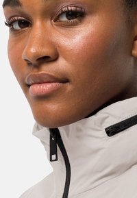 Lichte, crèmekleurige jas met een hoge kraag, voorzien van zwarte ritssluitingen en accenten, een gladde textuur en een minimalistisch ontwerp.