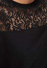 Top negro con intrincado panel de encaje floral sobre el pecho y el escote, mostrando piel a través de los patrones de encaje.