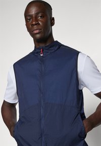 Un jeune homme portant un gilet sans manches bleu marine zippé par-dessus une chemise blanche à manches courtes, les mains dans les poches du gilet, regardant droit devant lui.