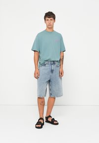 Light teal t-shirt with pink text, paired with faded denim shorts and black sandals. Simple design, relaxed fit, and casual style.