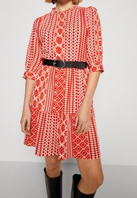 Robe rouge à motifs avec un devant boutonné, des manches 3/4 et un ourlet évasé, cintrée à la taille par une ceinture noire. Texture de tissu lisse.