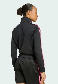 Donna con i capelli raccolti in un bun, che indossa una giacca sportiva nera e pantaloni corti con strisce rosa, in piedi di lato contro uno sfondo semplice.