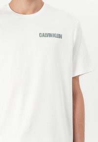 White cotton t-shirt featuring a crew neck and short sleeves, with a grey "CALVIN KLEIN" logo printed on the left chest.