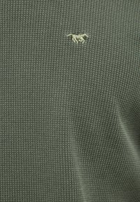 Grønt tekstureret stof med en lille, broderet logo i en lysere grøn nuance, der viser et ternet mønster og en blød finish.