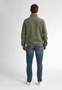 Giacca softshell verde oliva con colletto, yoke posteriore e cuciture piatte, abbinata a jeans in denim blu medio e sneakers bianche.