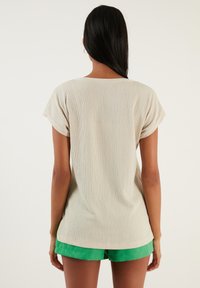 Haut beige texturé à manches courtes avec une coupe décontractée, doté d'un col ras du cou. Porté avec un short vert, montrant la vue arrière du shirt.