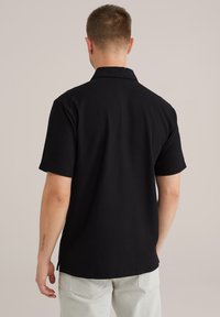 Črna kratkosrajčna srajca s kratkimi rokavi in teksturiranim rebrastim vzorcem, z ovratnikom in ravnim robom. Ima stranske razpoke in sproščen kroj.