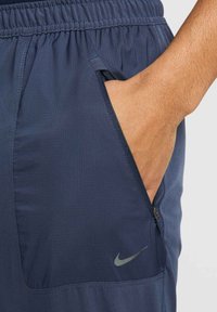 Granatowe szorty sportowe z elastycznym pasem, z zamkniętą kieszenią po boku oraz srebrnym logo Nike z przodu po lewej stronie. Teksturowana tkanina.