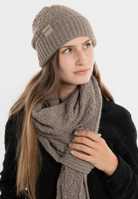 Khaki gebreide beanie met een gemêleerd afwerking en een merklabel, gedragen met een bijpassende dikke sjaal met een vergelijkbare textuur. Zwart geribbelde trui.