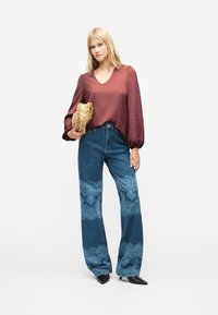 Γυναίκα με μπλούζα σε σκουριά απόχρωση και τζιν παντελόνι με φαρδιές μπατζάκια και δαντελωτά σχέδια, κρατώντας μια μπεζ τσάντα φάκελο με βολάν, στέκεται μπροστά σε λευκό φόντο.