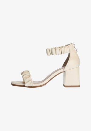 Sandalia de tacón beige con tacón cuadrado, tira con volantes alrededor del tobillo y una tira lisa sobre los dedos, con un diseño minimalista.