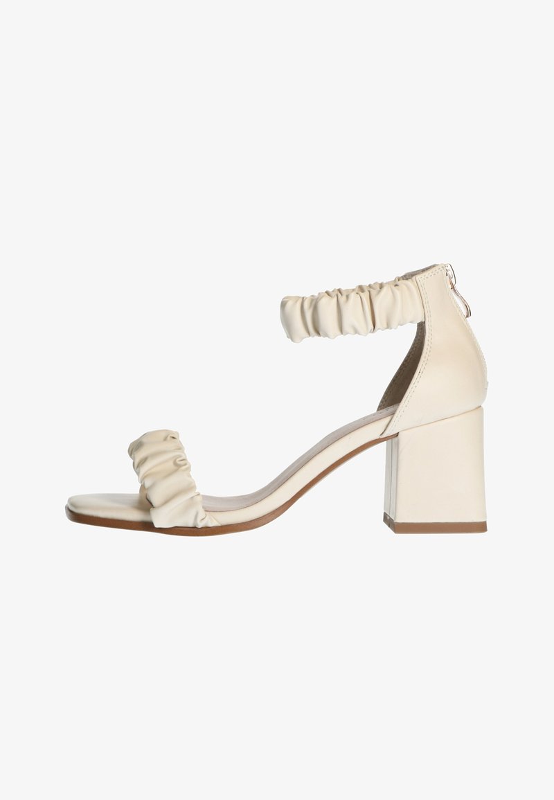 Sandale à talon beige avec talon bloc, bride froufroutante à la cheville et une bride lisse sur les orteils, présentant un design minimaliste.