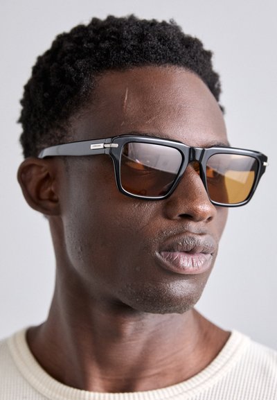 Lunettes de soleil carrées noires avec des verres teintés ambre, dotées de montures noires élégantes et d'accents argentés sur les branches. La peau texturée est visible.