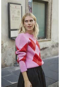 Różowy i czerwony sweter z dzianiny w kratę typu argyle z prążkowanymi mankietami, zestawiony z czarną spódnicą. W tle znajduje się kamienny mur i tablica z oznaczeniem drogi.