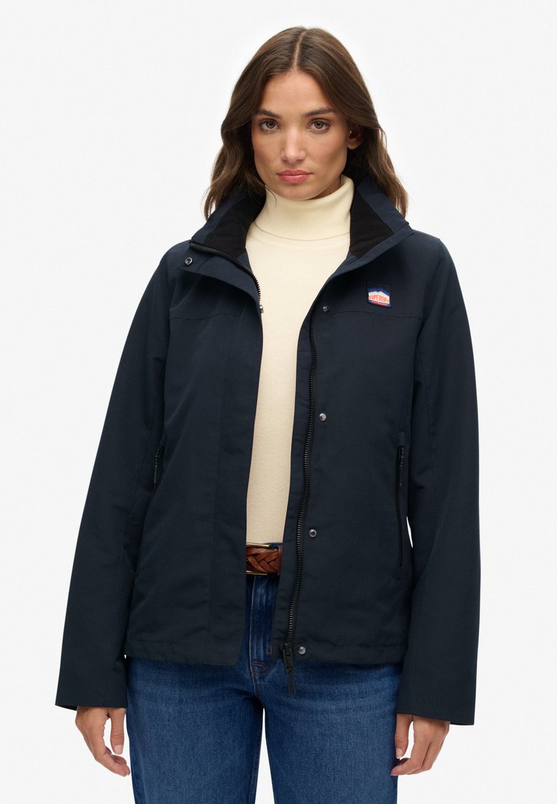 Marineblauwe waterdichte jas met een rits aan de voorkant, een opgeslagen kraag en een logopatch. Heeft zakken en een aansluitend ontwerp, gedragen over een lichte coltrui.
