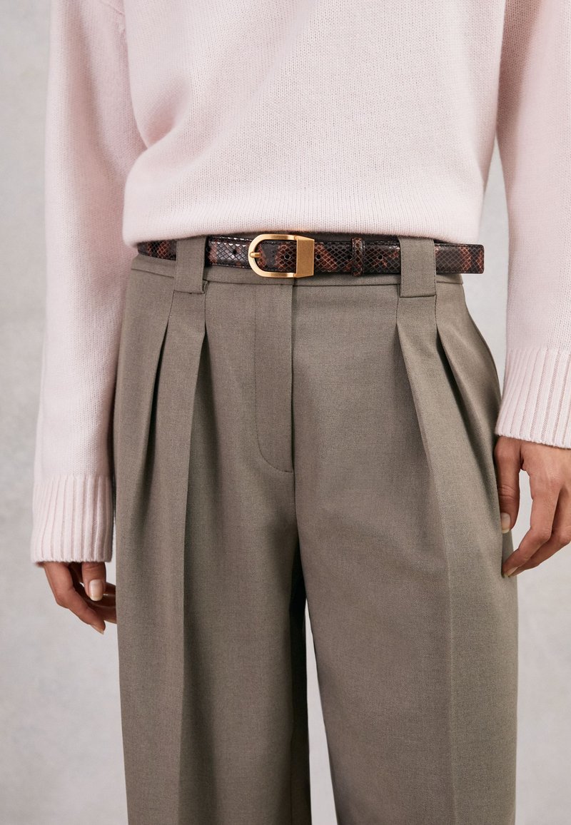 Frau trägt beige plissierte Hose mit braunem Schlangenprint-Gürtel und weichem rosa Strickpullover, steht vor neutralem Hintergrund.