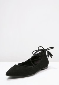 Bailarinas de ante negro con punta afilada, detalles de cordones y acentos de borlas. Presenta una suela plana con un acabado suave.