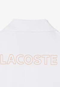Weißer Polokragen mit dem Schriftzug "LACOSTE" in großen, dünnen, orange umrandeten Buchstaben über dem oberen Rücken.