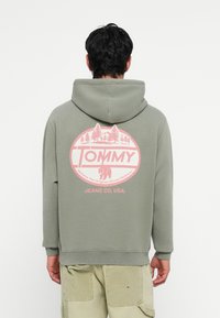 Groene hoodie met een roze cirkelvormige graphic met bomen en een beer. Tekst leest "TOMMY" en "JEANS CO, USA" onder het ontwerp.