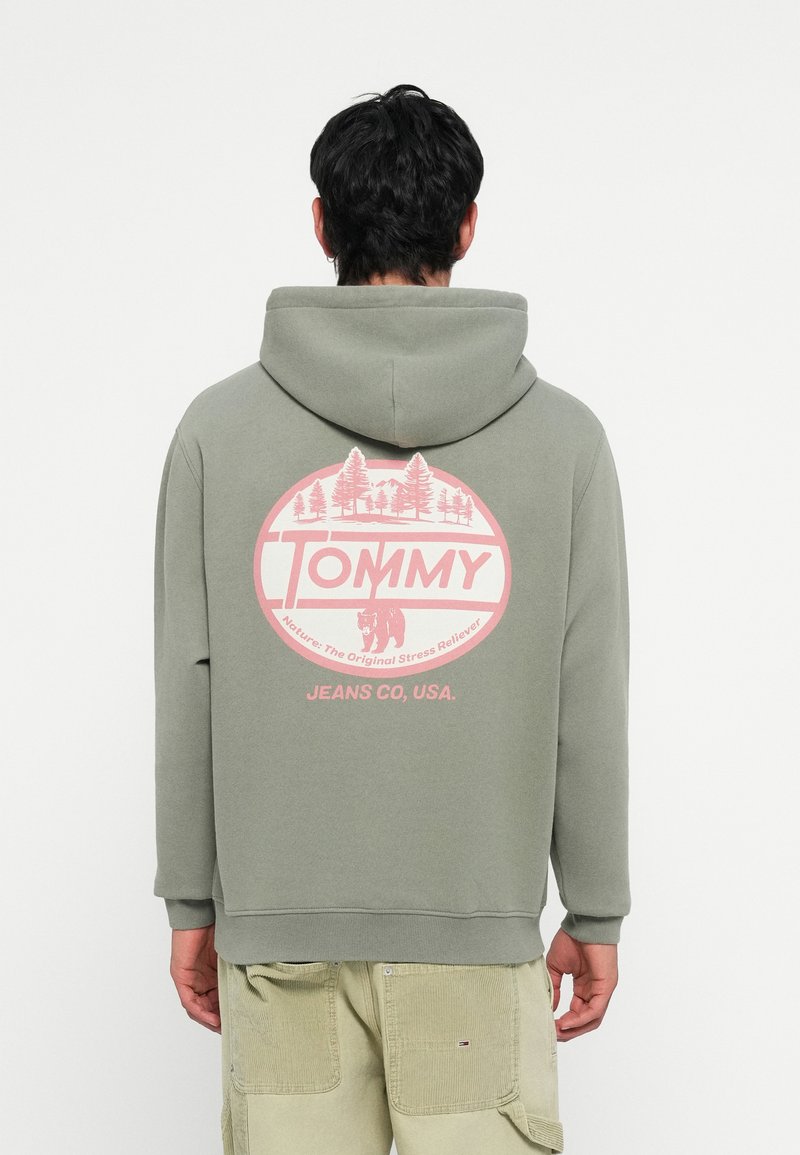 Groene hoodie met een roze cirkelvormige graphic met bomen en een beer. Tekst leest "TOMMY" en "JEANS CO, USA" onder het ontwerp.