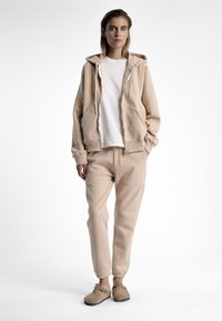 Hoodie beige avec poches avant, associé à un jogging assorti et des chaussures slip-on beige décontractées. Texture lisse, coupe décontractée et design minimaliste.