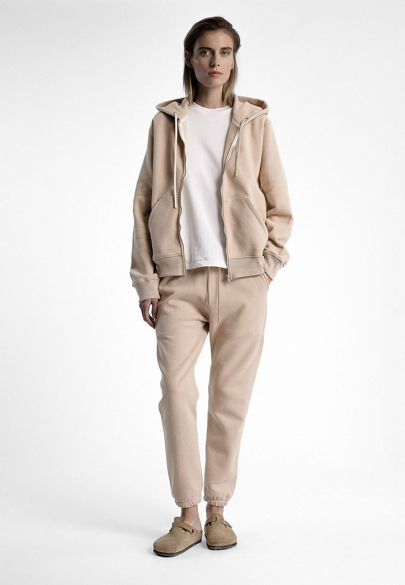 Hoodie beige avec poches avant, associé à un jogging assorti et des chaussures slip-on beige décontractées. Texture lisse, coupe décontractée et design minimaliste.