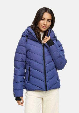 Femme portant une doudoune bleue avec capuche, fermeture éclair noire et pantalon blanc, se tenant devant un fond clair.