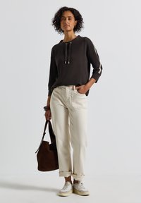 Dunkelbrauner Hoodie mit Kordeln, cremefarbene, schmal zulaufende Hose und graue Sneakers. Hält eine braune Wildledertasche. Lockiges Haar, neutraler Hintergrund.