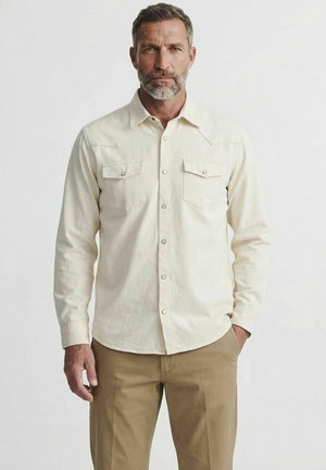 Mann mit grauem Bart, der ein cremefarbenes langärmeliges Hemd mit Klappentaschen und Khakihosen vor einem schlichten hellen Hintergrund trägt.