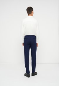 Camicia bianca a maniche lunghe, pantaloni blu navy, scarpe nere. I pantaloni presentano passanti per cintura e tasche posteriori, vestibilità slim con una texture liscia.