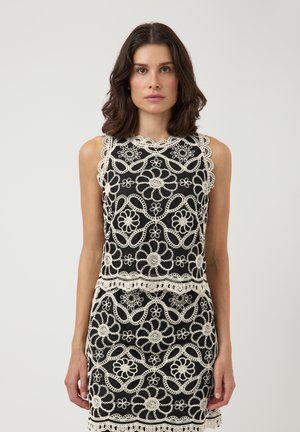 Mujer con conjunto negro de top sin mangas y falda con bordado de encaje floral blanco, de pie frente a un fondo claro y sencillo.