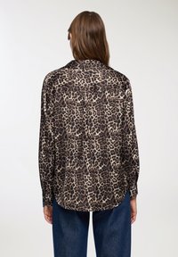 Bluzka w panterkę z lekkiego materiału, o luźnym kroju, z długimi rękawami i zaokrąglonym dołem. Dominują ciemne brązy i neutralne odcienie.