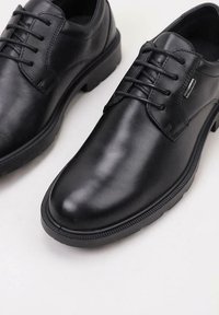 Chaussures de ville noires en cuir lacées avec des bouts ronds et des semelles en caoutchouc texturé sur un fond clair.