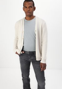 Cárdigan de punto beige claro con puños acanalados, usado sobre una camiseta gris claro y combinado con jeans gris oscuro, con un sencillo cierre de botones.