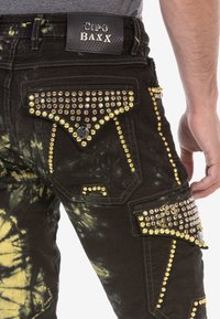 Zbliżenie czarno-żółtych spodni w stylu tie-dye z złotymi i brązowymi ćwiekami na kieszeniach i szwach, noszonych przez osobę z widoczną ręką.