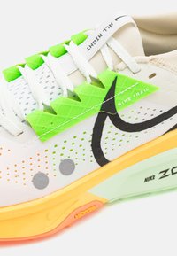 Les chaussures Nike Trail présentent une tige beige clair avec des accents verts, des lacets blancs et une semelle texturée aux détails orange et jaune.