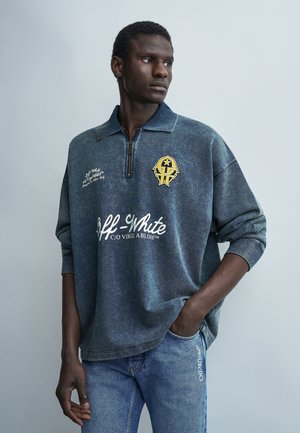 Νεαρός άντρας που φοράει φθαρμένο μπλε πουλόβερ Off-White με κίτρινο έμβλημα και τζιν με λεπτομέρεια τσέπης με λογότυπο Off-White, κοιτάζοντας πλάγια.