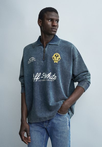 Jeune homme portant un pull-over bleu délavé Off-White avec un emblème jaune et un jean avec un détail de poche à logo Off-White, regardant sur le côté.