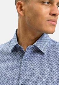 Blå mönstrad skjorta med krage, som har en geometrisk design, kontrasterande knappar och en mjuk tygstruktur.