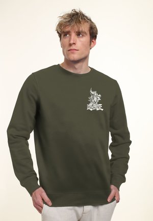 Young man with tousled blond hair wearing an olive green Yu-Gi-Oh! sweatshirt and light-colored pants, looking slightly to the side.
