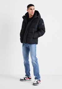 Giacca a vento nera con cappuccio, caratterizzata da un design trapuntato, chiusura con zip e tasche laterali. Abbinata a jeans azzurri e sneakers nere-bianco-rosse.