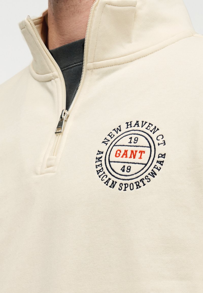 Felpa beige con mezza cerniera, caratterizzata da un logo circolare ricamato con la scritta "GANT" in arancione, insieme a "New Haven CT" e "American Sportswear".