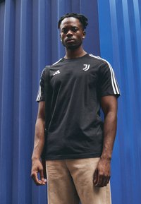 Sort atletisk t-shirt med hvide striber, med et logo på brystet. Modellen bærer khaki bukser og holder en skuldertaske. Blå baggrund.