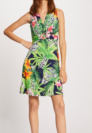 Woman wearing sleeveless V-neck dress with tropical floral and leopard print, belted at waist with round buckle, standing with hand on hip.