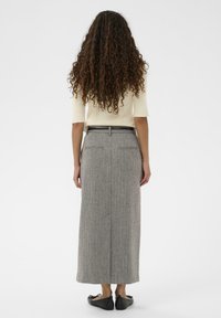 Jupe maxi en tweed gris avec une texture subtile, dotée de poches arrière et d'une ceinture noire élégante. Modèle portant des chaussures plates noires.