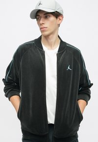 Casaco bomber de veludo preto com fecho éclair, apresentando detalhes em azul claro e um logótipo Jumpman; combinado com uma t-shirt branca simples.