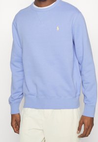Světle modrý crewneck svetr vyrobený z měkké tkaniny, s žebrovanými manžetami a lemem, se malým žlutým logem na hrudi.