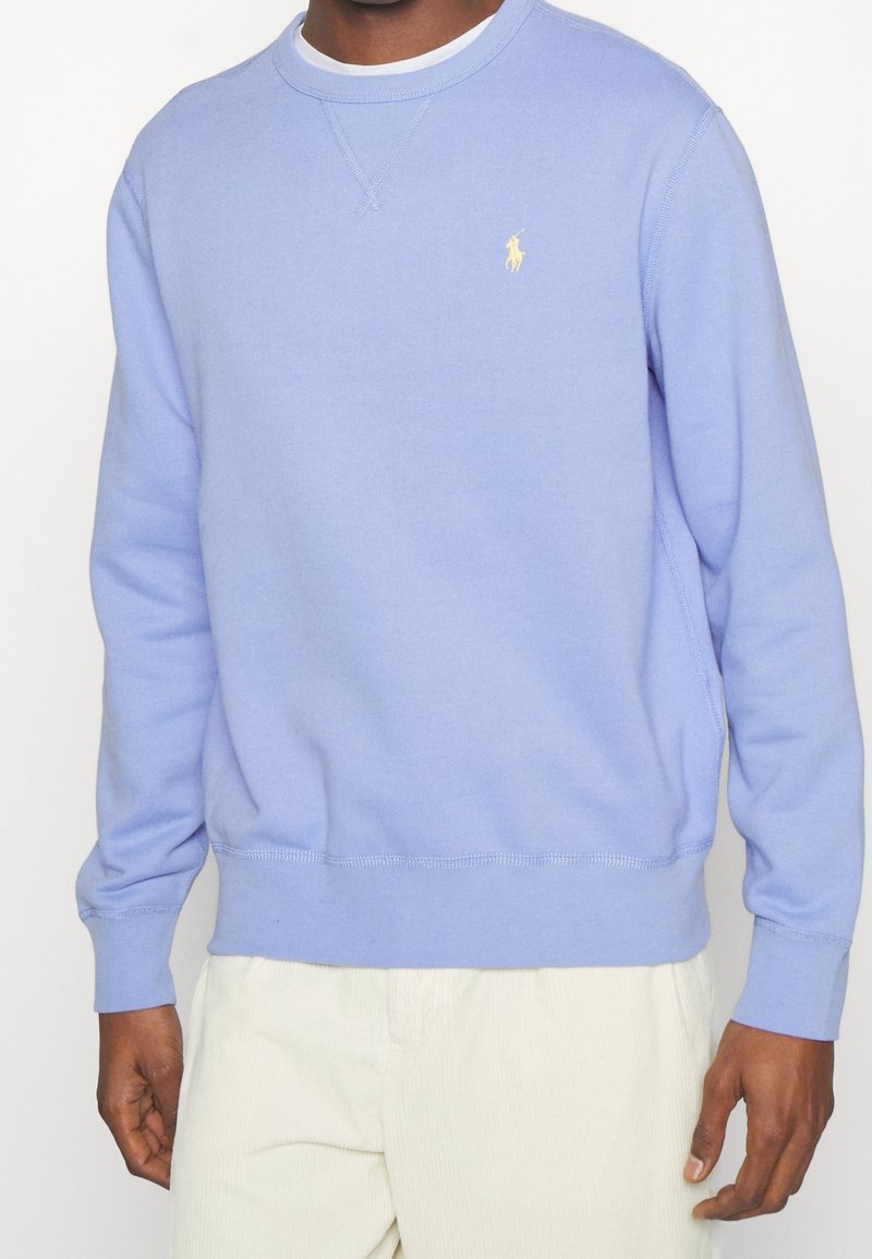 Světle modrý crewneck svetr vyrobený z měkké tkaniny, s žebrovanými manžetami a lemem, se malým žlutým logem na hrudi.