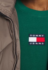 Grön bomullströja med en rektangulär marinblå lapp som har "TOMMY JEANS" och ett rödvitt horisontellt randmönster. Brun puffjacka delvis synlig.